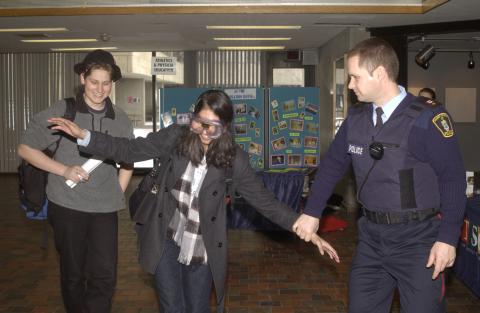 Campus Police Officer Demonstrates Sobriety Test with Student Wearing Impairment Goggles, Graditude 0T2 Week Event in the Meeting Place