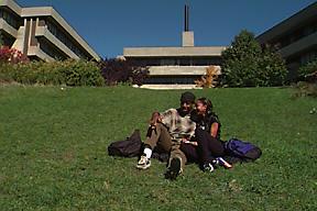 Two Students Sit on Grassy Slope below H-Wing Patio, Andrews Buildings Visible in Background