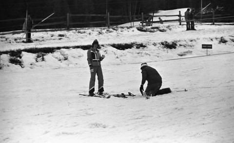 One Student Standing While Another Puts on Skis
